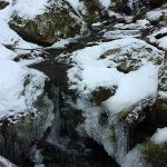 Stream surrounded by snow-covered ground and rocks, frozen water at edges of stream