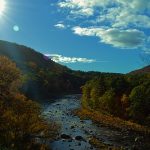 River surrounded by autumn trees and hills, blue sky with cloud and bright sun in upper left