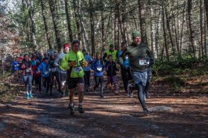 A large group of people running on the trail at the Chesterfield Gorge, wearing race bibs and dressed warmly. Sunlight shining through trees in the background.