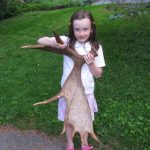 Young white girl standing in front of a lawn, holding a large moose antler that is nearly as tall as she is.