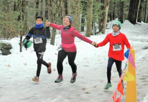 A woman runs through the finish line holding the hands of two children. Ice and snow cover the ground and trees are in the background