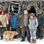 Eight people and a dog pose in front of a structure made from branches and twigs