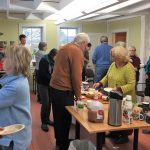 People standing around a table, helping themselves to pie and beverages, others in the room talk and look at pictures on the walls