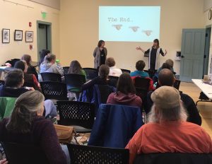 The audience is watching two women speak about their books. Behind them is a slide show with an image indicating that they are at the end of their presentation.