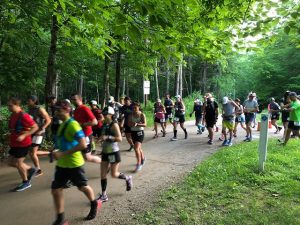 A crowd of runners take off from the starting line on a trail. Green trees are in the background.