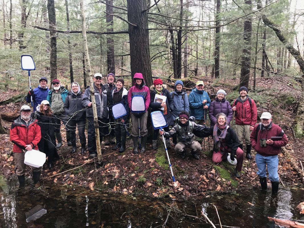 A couple dozen people pose for a photo in the woods behind a vernal pool