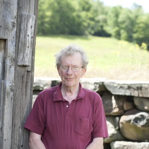 Wil Hastings, wearing glasses and a maroon shirt, is seen from the torso up. He is sitting in front of a stone wall, with a sunlit field behind him.