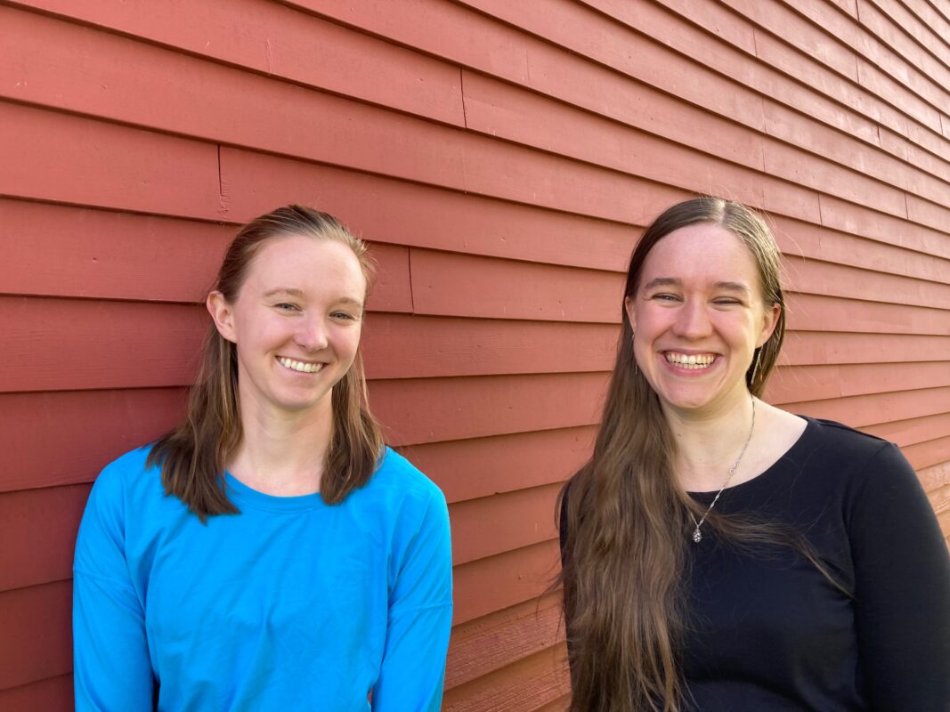 Audrey and Sarah shown from the mid-torso up, looking into the camera and smiling. Behind them is the outside wall of a red barn.