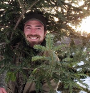 Michael Madole's face is visible, looking into the camera and smiling, peeking out from between the branches of a fir tree