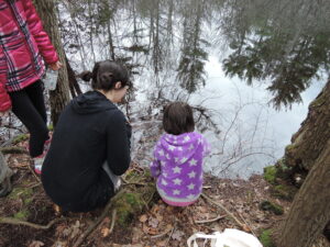 An adult and child squat at the edge of a vernal pool, looking into the water