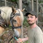 Michael, seen from the waist up, is wearing a brown hat and green shirt, and is looking into the camera and smiling. He is holding the reins of his tan horse Hazel, whose head with a blonde mane is next to him. Behind them are some small tree trunks and a rock wall.