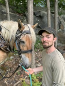 Michael, seen from the waist up, is wearing a brown hat and green shirt, and is looking into the camera and smiling. He is holding the reins of his tan horse Hazel, whose head with a blonde mane is next to him. Behind them are some small tree trunks and a rock wall.