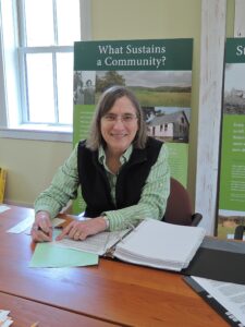 Suzy Kulik, sitting at a table with binders spread around her. She is looking into the camera and smiling.