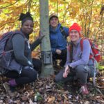 Tiffany, Tim, and Sarah, wearing backpacks and dressed for fall New England weather, pose for a photo squatted in the woods near the base of a birch tree where a wildlife camera is secured. Behind them are autumn leaves and rocks.