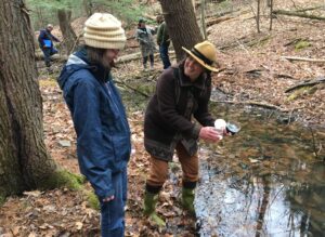 Two people stand in at the edge of a vernal pool in a leaf covered forest in early spring. One of them holds various jars in their hands with samples collected from the pool, looking back and smiling at the other person. Three more people in the background are visible on the far edge of the pool.