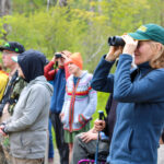 A group of people, some of whom are looking through binoculars, standing and seated at the edge of a beaver meadow.
