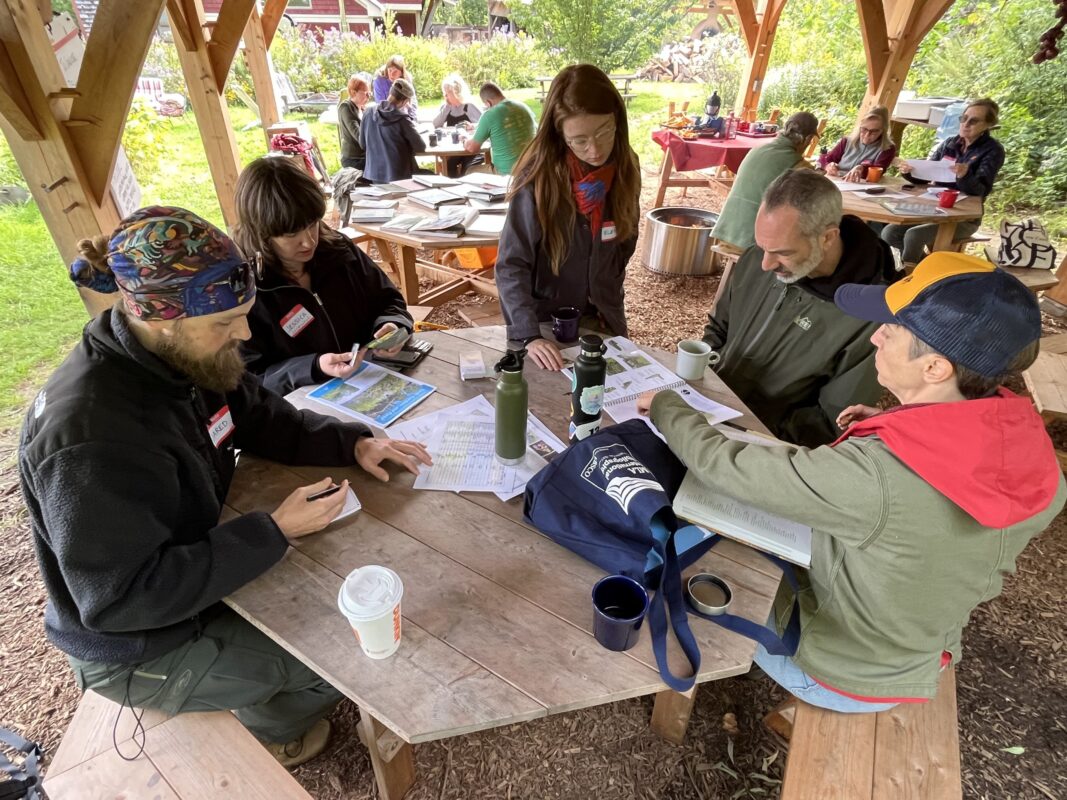 A wide shot of the whole group sitting underneath the pavilion at picnic tables. In the foreground, four people seated at the table are looking at notebooks and planting plans while Elena, standing, talks with them.