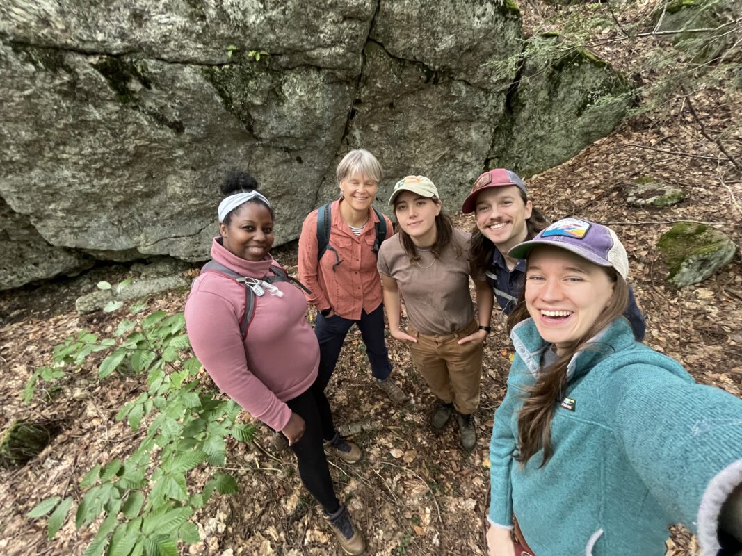 A fish-eye view of five people dressed in hiking clothes, standing shoulder to shoulder and smiling, with a rock ledge in the background.