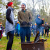 Two warmly dressed people smile ateach other as they warm their hands over a fire pit.