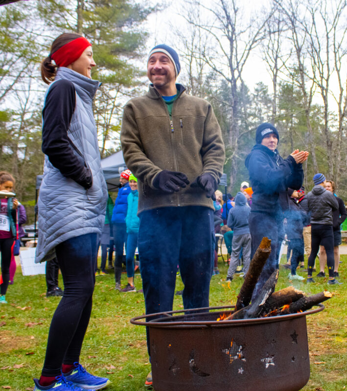 Two people smile at each other as they gather around a fire pit to stay warm. A crowd is visible in the background on the overcast day.