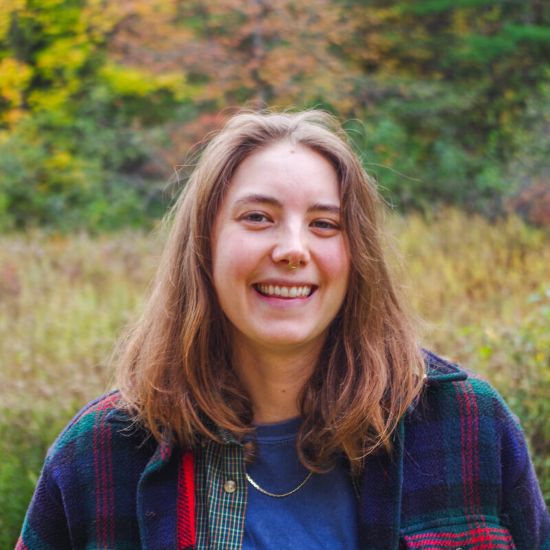 Seen from the shoulders up in warm fall clothing in front of blurred fall foliage, Mariel smiles into the camera.