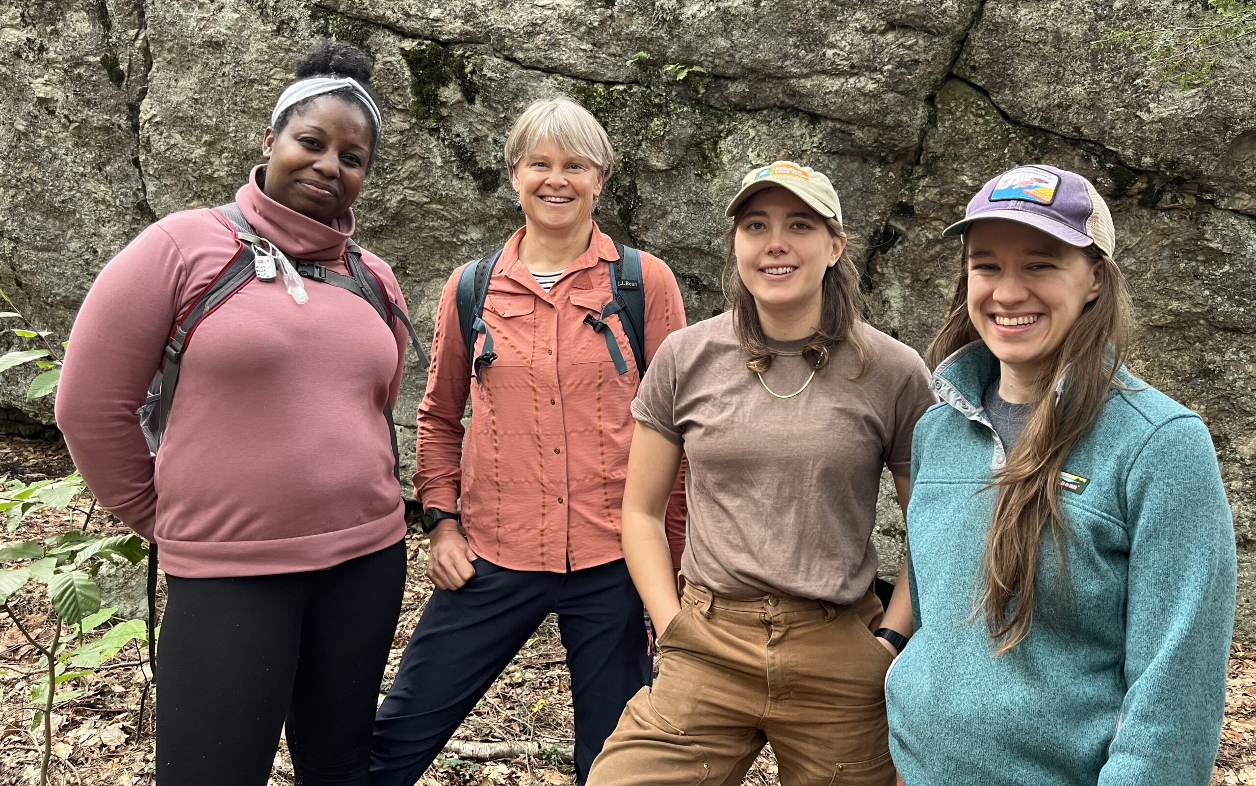 Standing in a row in front of a rock face, Tiffany, Sally, Mariel, and Sarah are smiling and dressed in fall weather hiking clothes.