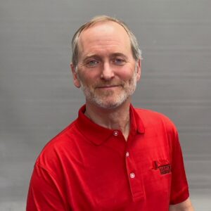 A professional-style headshot of Tad, who is wearing a red polo shirt and standing in front of a metallic gray background.