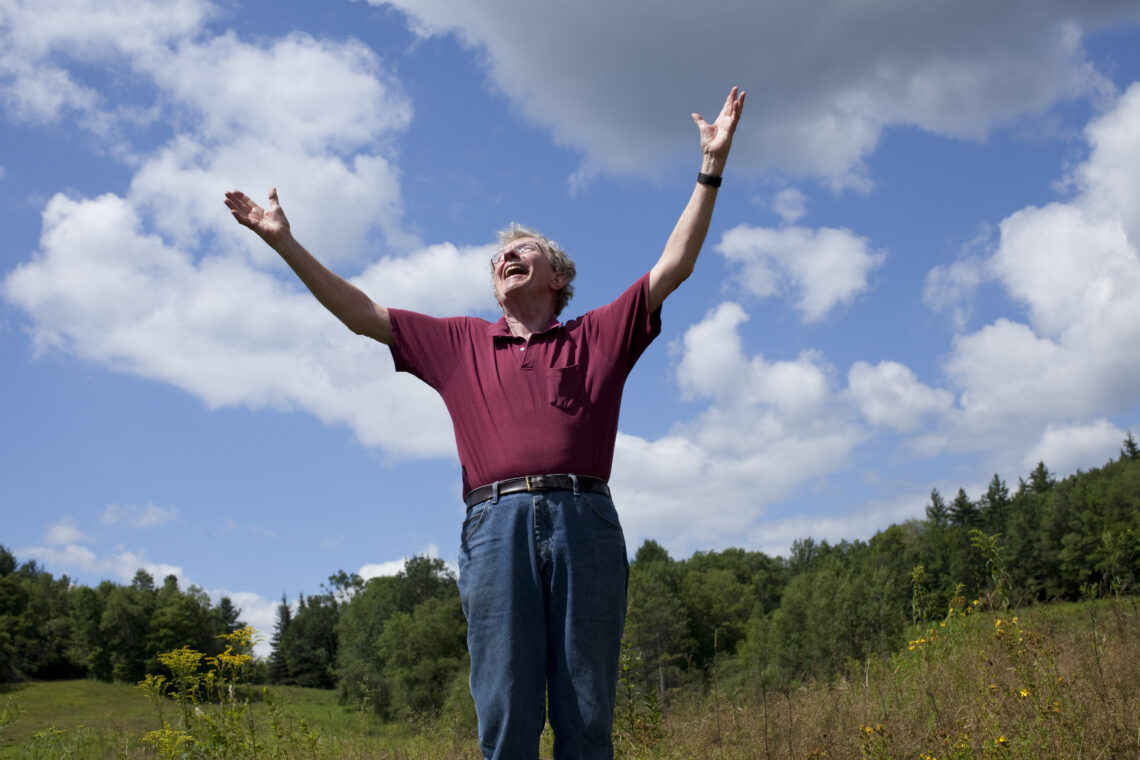 Wil Hastings, standing in a field, smiles and throws his arms joyously into the air.