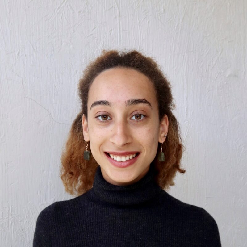 Juliette, who is wearing a black turtleneck and dangle earrings, is seen from the shoulders up smiling into the camera. They are standing in front of a textured white wall with their curly hair pulled back.
