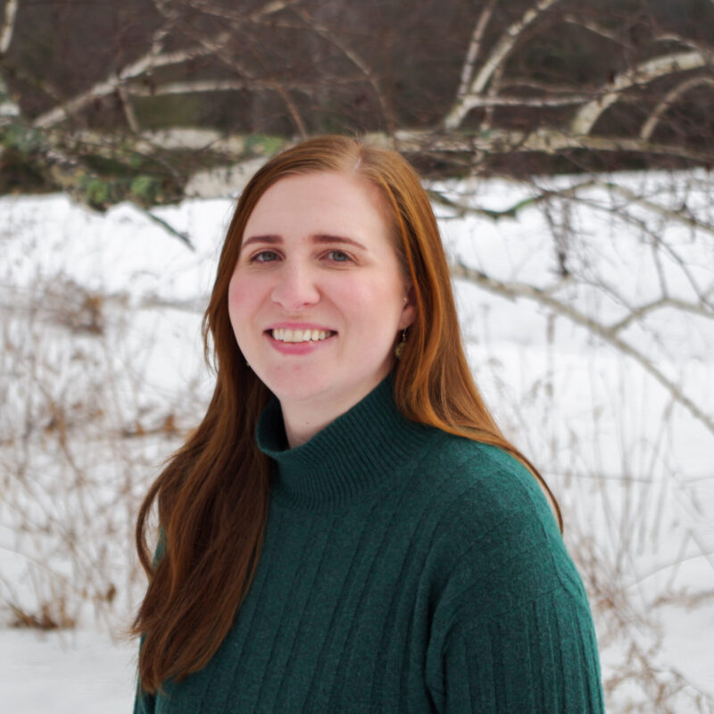 Alli is seen from the torso up wearing a green sweater and standing in front of a birch branch and a snowy field. She is smiling towards the camera.