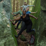 Tiffany is in the foreground of a group of HLT people including Sally, Sarah, and Tim. Using their hands and feet to press against either side of a giant, moss-covered split boulder, they are suspending themselves above the ground.
