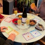 Bingo cards, snacks, and wineglasses on a round table, with people's hands visible on the edges.