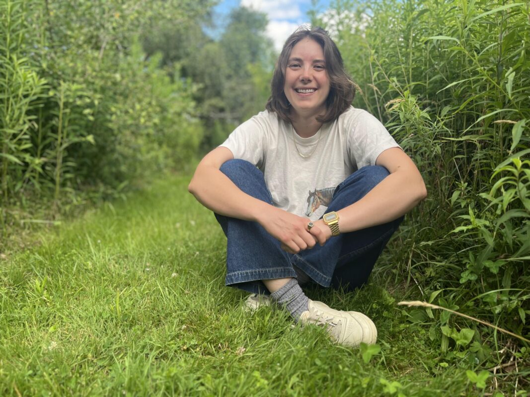 Mariel, smiling sitting cross-legged on a grassy mown path through a dense wildflower meadow. A slice of blue sky is seen through the meadow.