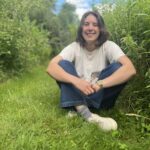 Mariel, smiling sitting cross-legged on a grassy mown path through a dense wildflower meadow. A slice of blue sky is seen through the meadow.