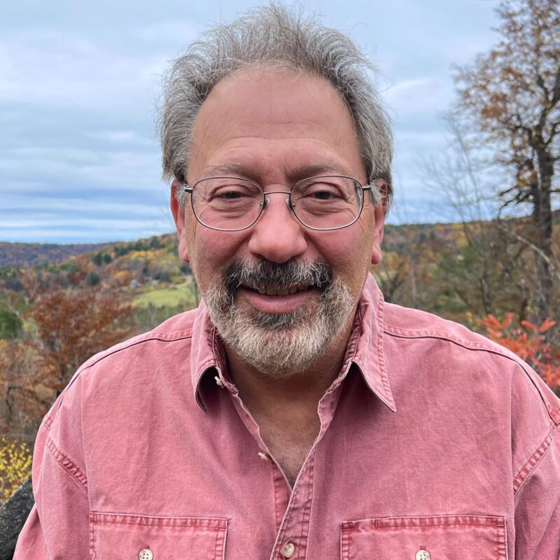 Gary, who is wearing glasses and a faded red shirt and smiling into the camera. Cloudy skies and autumn hills are behind him.