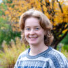 A headshot style photo of Calla, who is wearing a gray sweater and smiling in front of a yellow-leaved tree.