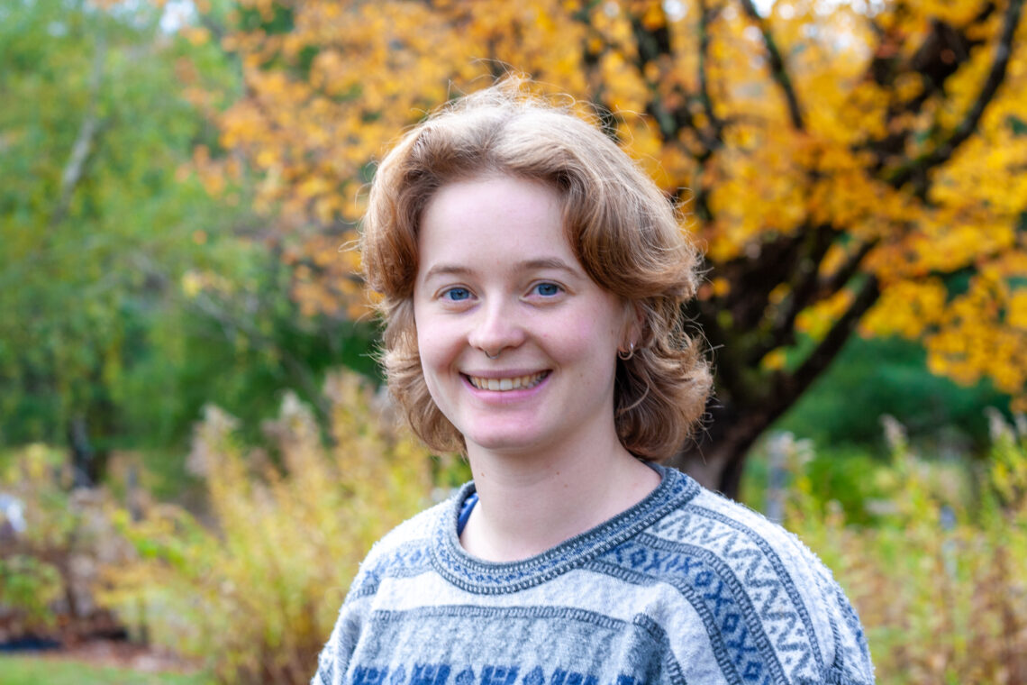 A headshot style photo of Calla, who is wearing a gray sweater and smiling in front of a yellow-leaved tree.
