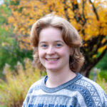 A headshot style photo of Calla, who is wearing a gray sweater and smiling in front of a yellow-leaved tree.