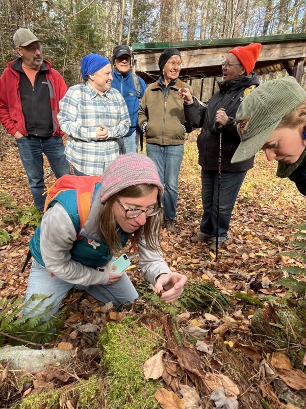 A group of seven warmly dressed people gathers around a forest floor, laughing together and investigating autumn mushrooms.