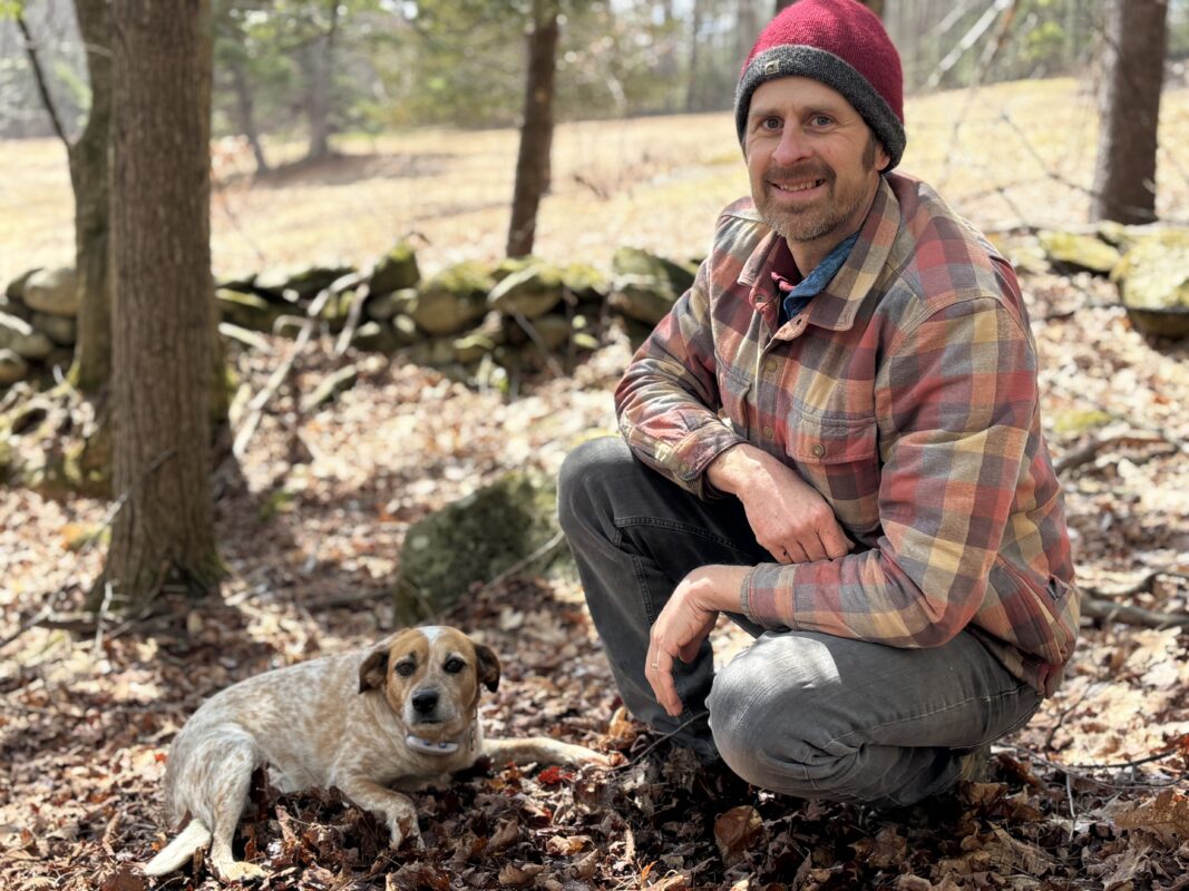A photo of James Thompson at the edge of a field kneeling and smiling with his dog.
