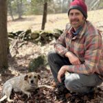 A photo of James Thompson at the edge of a field kneeling and smiling with his dog.