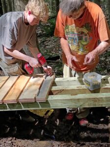 Two people work together to screw in planks onto a bridge. 
