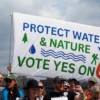 Banner that reads "Protect water & nature vote yes on 6" held above a crowd of people